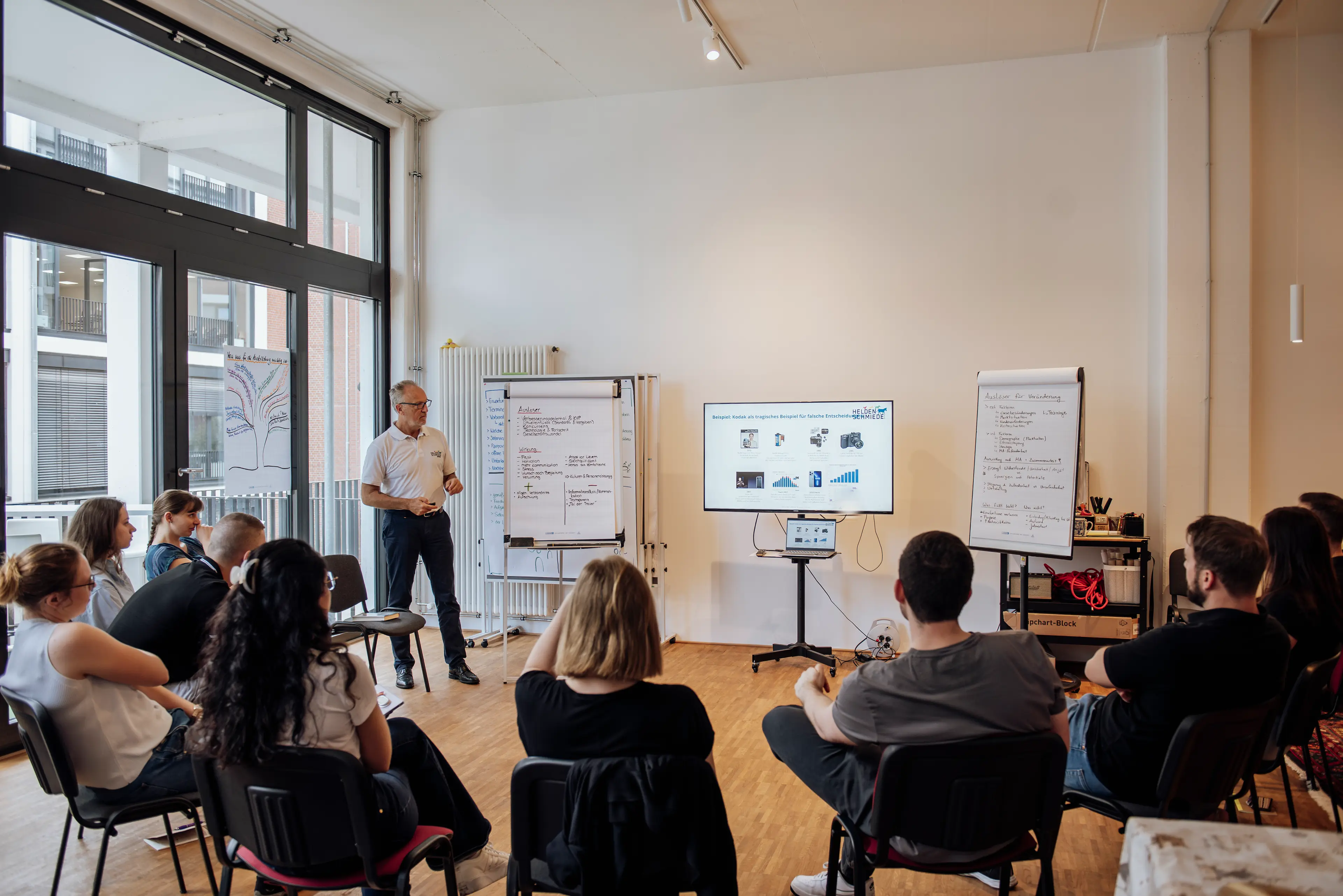 Armin Straub steht neben zwei Whiteboards und einem TV vor einer sitzenden Gruppe Seminarteilnehemden.