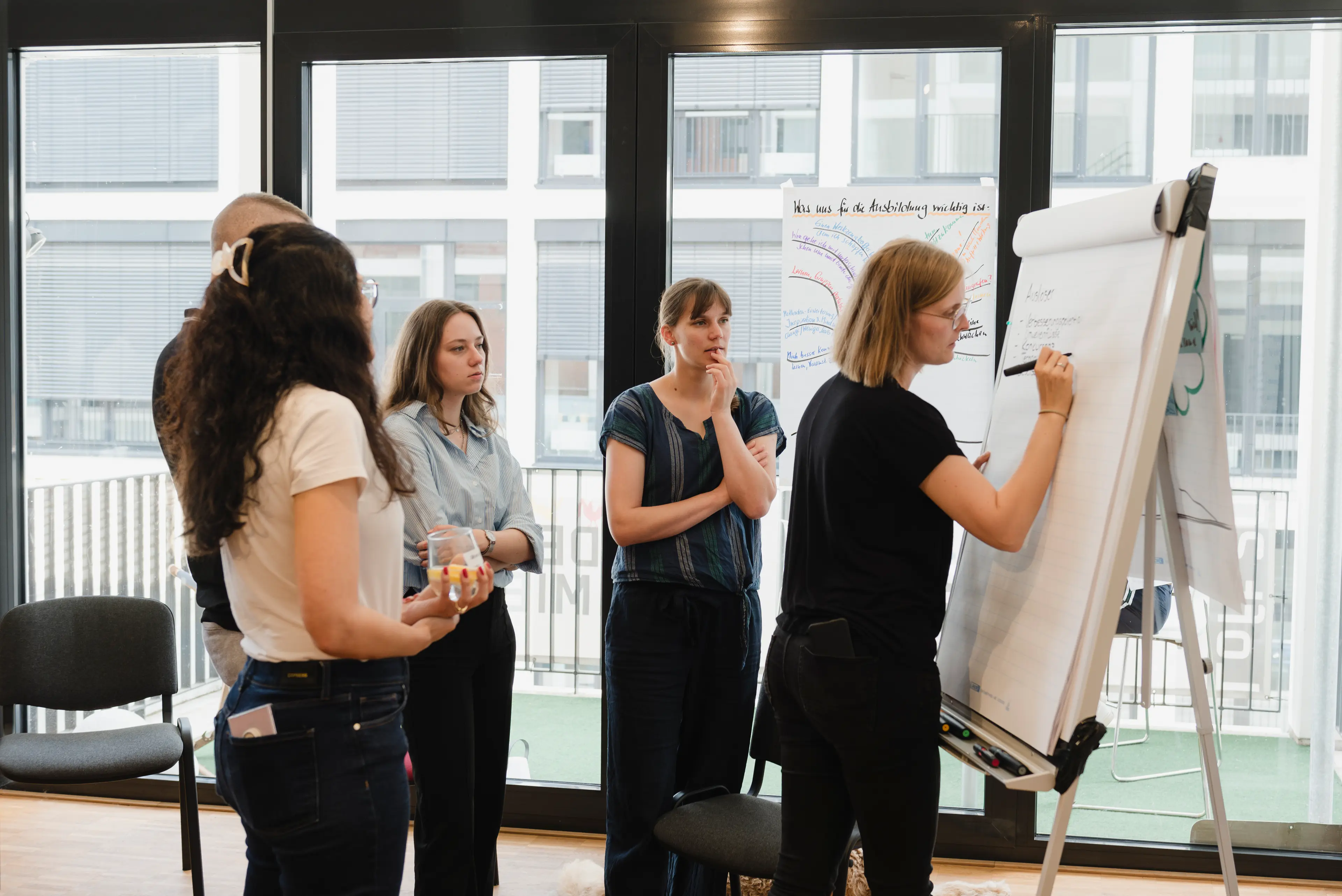 Fünf Seminarteilnehmende, die im Halbkreis vor einem Whiteboard stehen. Eine der Personen schreibt gerade etwas an das Whiteboard.